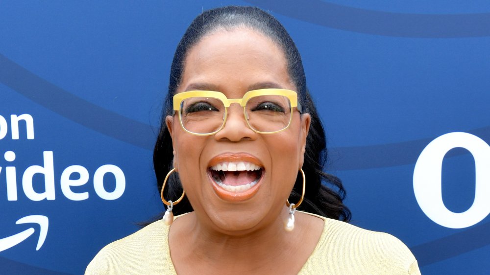 Oprah Winfrey in a yellow dress and glasses, laughing while looking straight at the camera