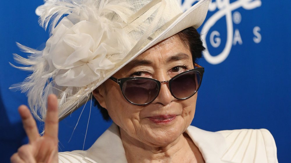 Yoko Ono in a white blazer, hat, and sunglasses, smirking and giving the peace sign