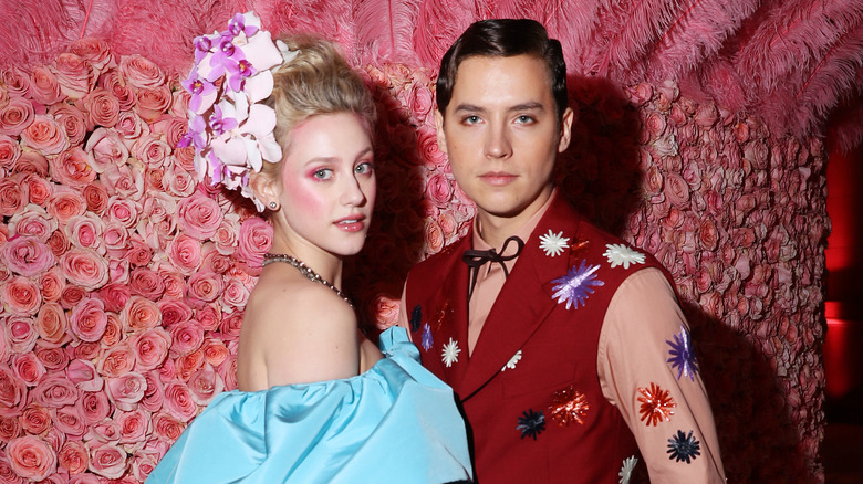 Lili Reinhart in a couture flowered wig and sleeveless blue gown with Cole Sprouse wearing a red floral vest and long sleeved shirt in front of a wall of flowers