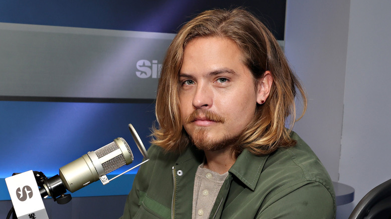 Dylan Sprouse with a microphone while wearing a green bomber jacket and tan button up shirt