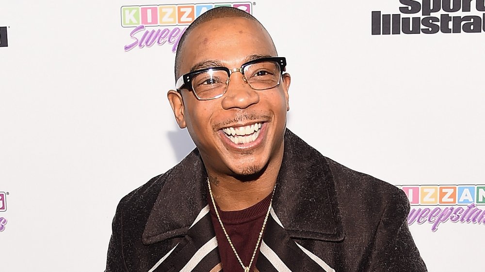 Ja Rule in a brown coat and black-rimmed glasses, smiling at a step-and-repeat