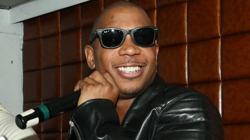 Ja Rule in a black leather jacket and black sunglasses, smiling while holding a mic