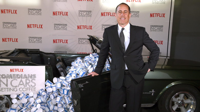 Jerry Seinfeld smiling next to car full of coffee cups and a sign for Comedians in Cars Getting Coffee