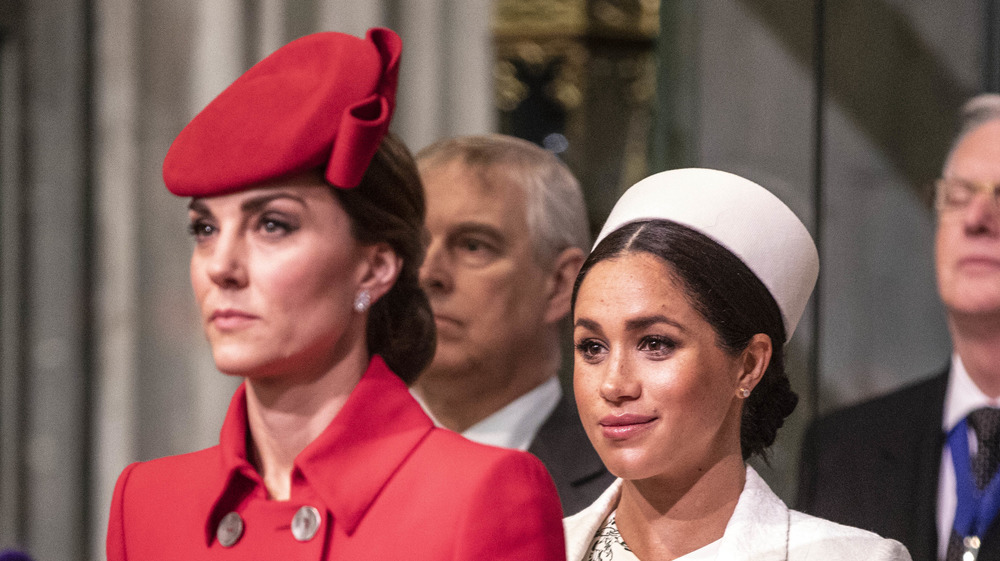 A stone-faced Kate Middleton standing in front of a smirking Meghan Markle