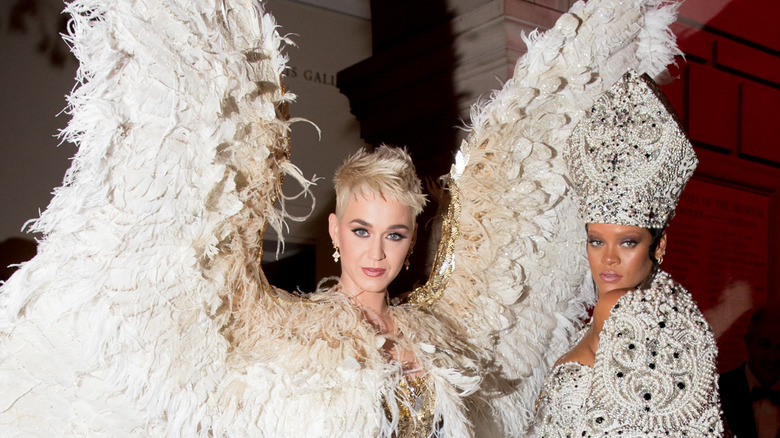 Katy Perry wearing large white feathers and Rihanna wearing a bejeweled pope's hat and jacket