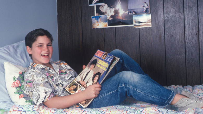 Joaquin Phoenix as a child wearing a newspaper-patterned button down shirt and jeans while lying on a bed