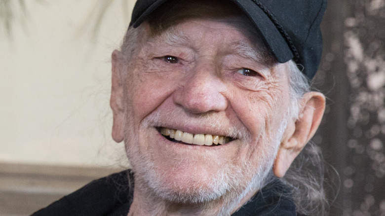 Willie Nelson smiles and wears a black baseball hat