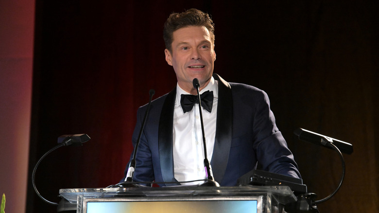 Ryan Seacrest speaks at the microphone wearing a tux