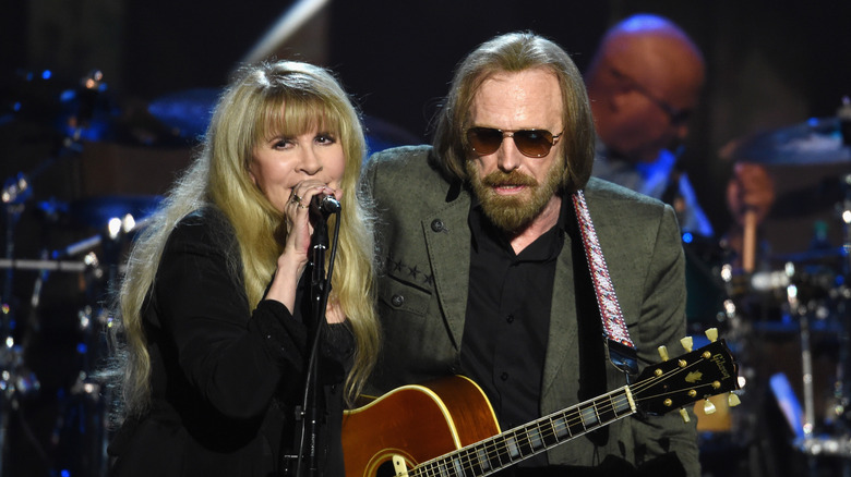 Stevie Nicks wearing a black jacket and dress singing with Tom Petty wearing a green-grey jacket and black shirt