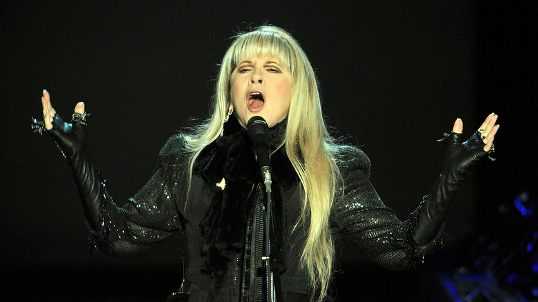 Stevie Nicks singing while wearing a black bedazzled leather coat and black leather fingerless gloves