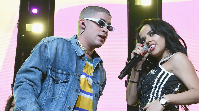 Bad Bunny wearing a denim jacket and white sunglasses performing with Becky G wearing a black and white sparkly leotard