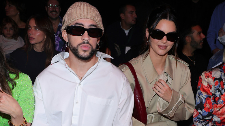 Bad Bunny in a white collared shirt and brown beanie with Kendall Jenner in a tan trenchcoat and black sunglasses