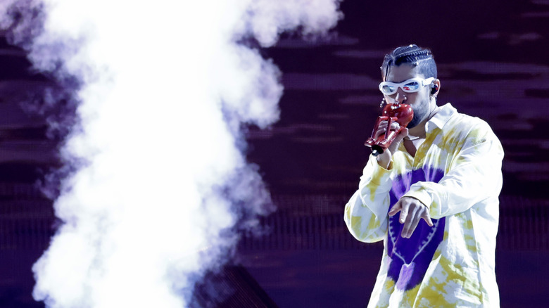 Bad Bunny performing on stage with a cloud of smoke while wearing a white, blue, and yellow long sleeved shirt