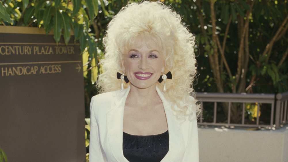 Dolly Parton smiling big in a white suit and black top in 1985 at age 39