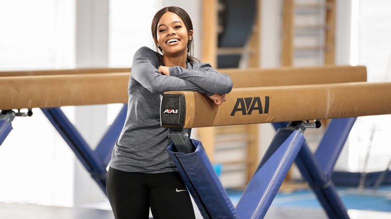 Gabby Douglas smiling and leaning against gymnastics beam