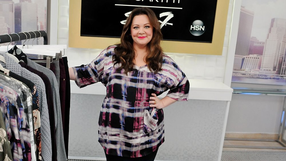 Melissa McCarthy posing next to a clothes rack for her Seven7 clothing line