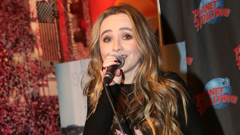 Sabrina Carpenter holding a microphone wearing a long sleeved black top