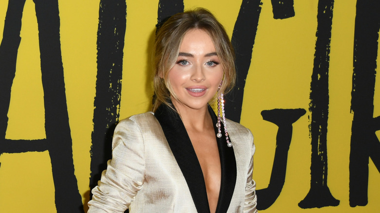 Sabrina Carpenter with dark blonde hair wearing a gold suit with black lapel and long dangling earrings