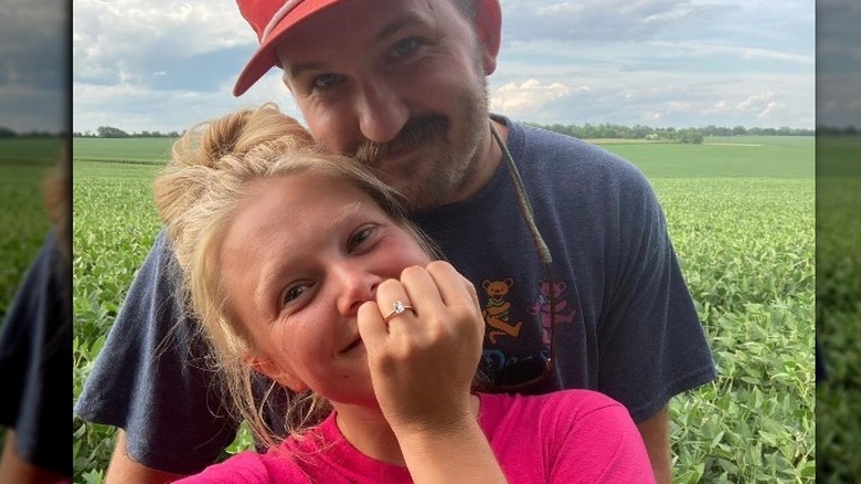 Hailey Whitters, smiling, no makeup, messy bun, showing off her engagement ring, selfie, Jake Gear behind her, smiling, wearing a hat