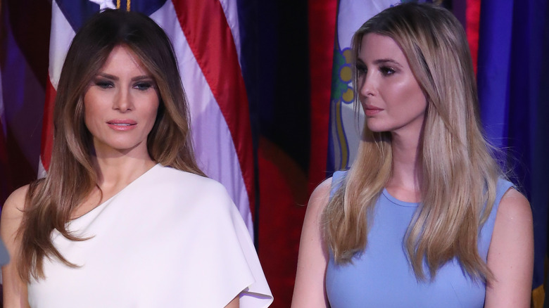 Melania Trump in a white shall dress, Ivanka Trump in a blue dress, standing next to each other with serious expressions