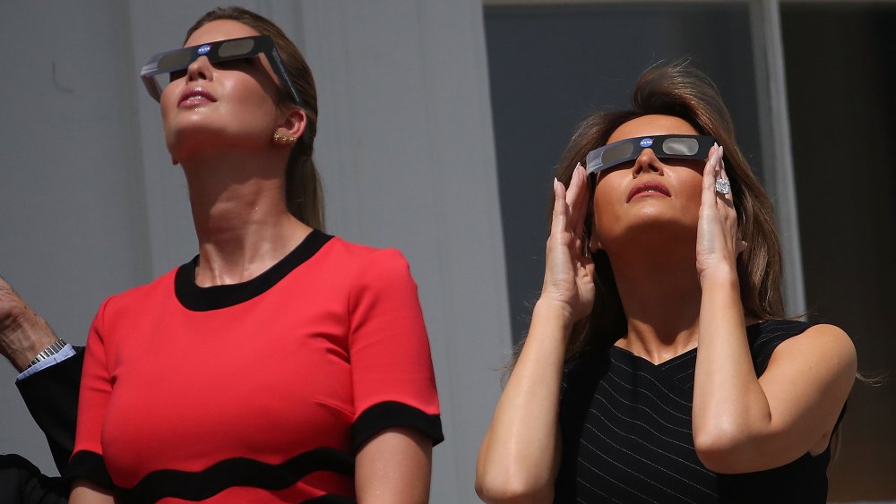 Ivanka Trump in a red-and-black dress, Melania Trump in a black pin-striped dress, both looking up and wearing glasses to watch an eclipse