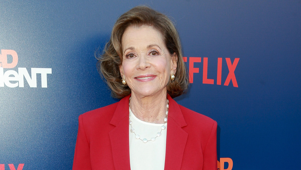 Jessica Walter wearing gold earrings and a red blazer smiles for a photo on the red carpet 