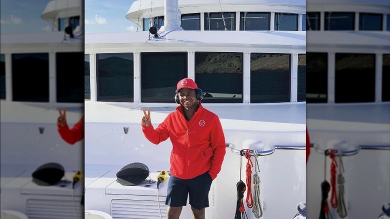 Mzi 'Zee' Dempers, smiling, giving the peace sign on the Yacht's deck, wearing a red hat and jacket 