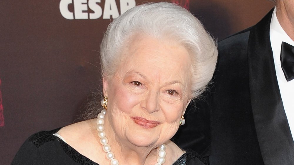 Olivia de Havilland in a black dress and pearls, with a small smile at a red carpet event
