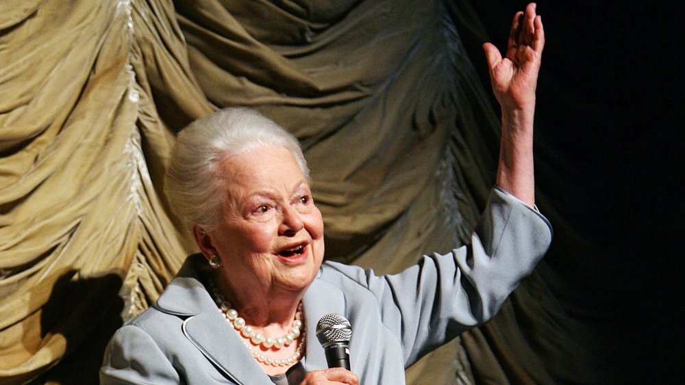 Olivia de Havilland in a blue outfit and pearls, speaking into a mic