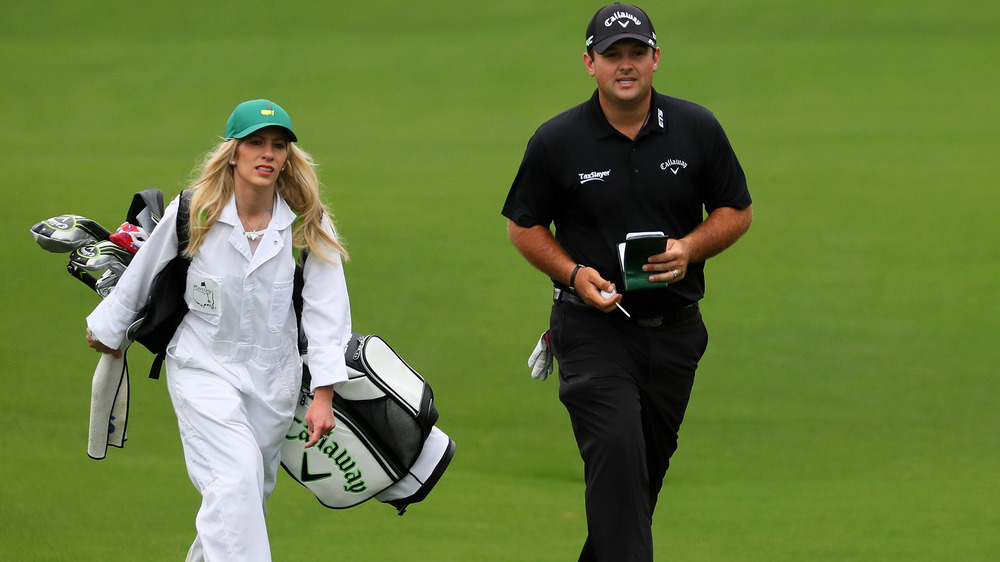 Justine Reed, working as caddie, wearing all white, wearing a hate, holding golf clubs; Patrick Reed, wearing all black and a hat, on the golf course