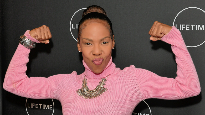 Drea Kelly flexes biceps in a pink turtleneck sweater.