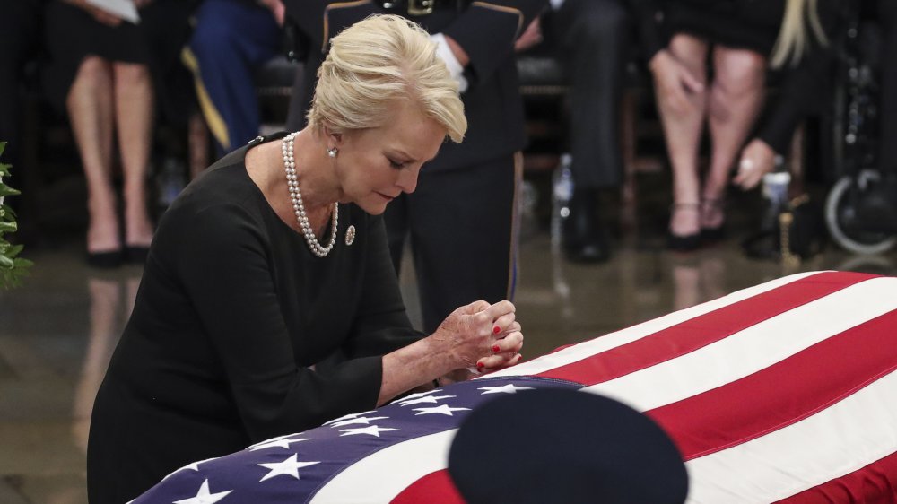 Cindy McCain wearing black and pearls, kneeling and praying over John McCain's casket