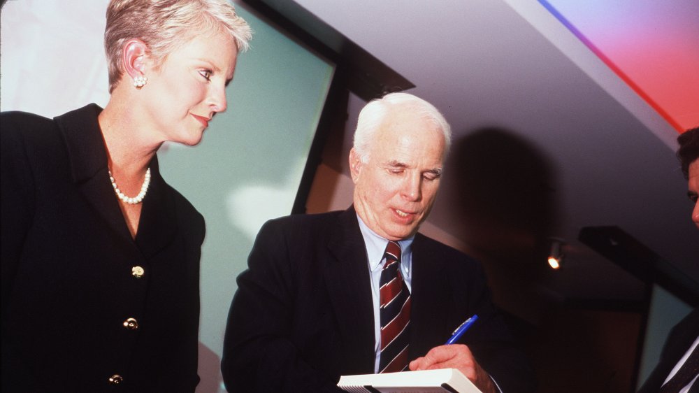 Cindy McCain in a black blazer dress and pearls, watching as husband John McCain signs a book in 1999