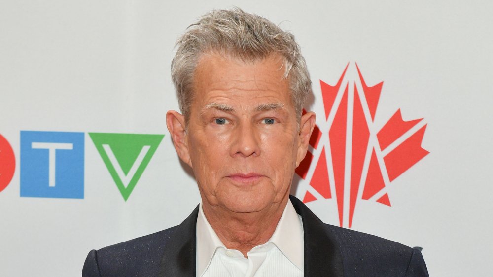 David Foster posing with neutral expression on a red carpet, wearing a black blazer and white dress shirt