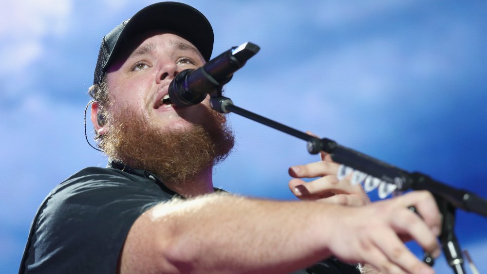 Luke Combs in a black shirt and cap, adjusting his mic on stage