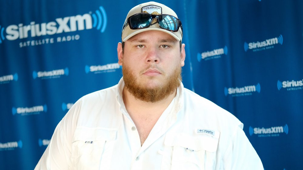 Luke Combs in a white button up, hat, and sunglasses, posing with a serious expression at a SirusXM event