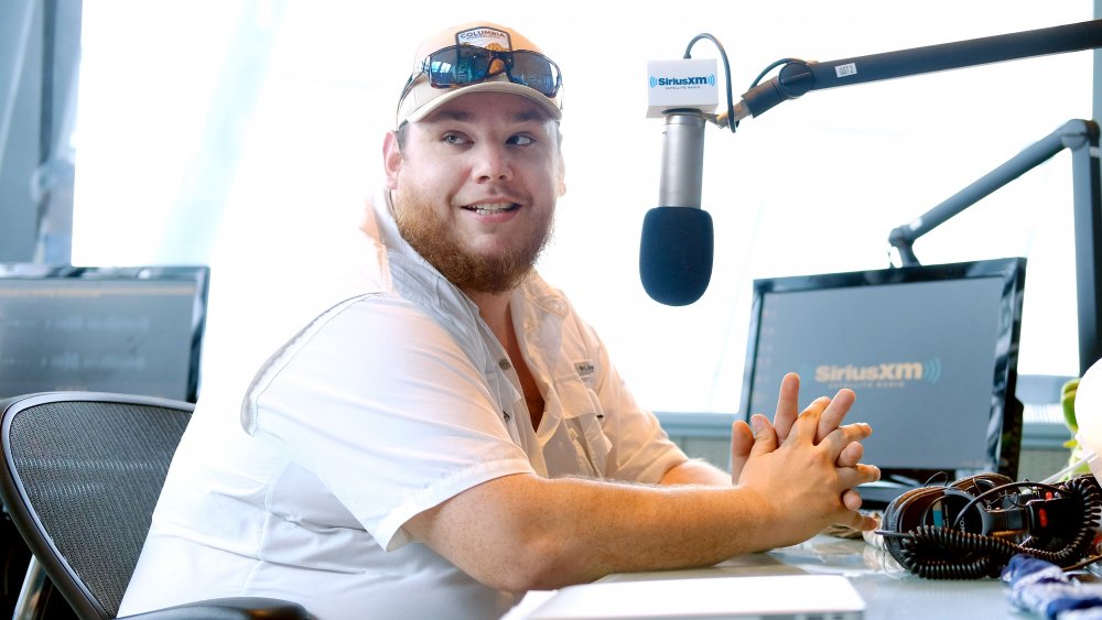 Luke Combs in a white shirt and hat, speaking on a SiriusXM show