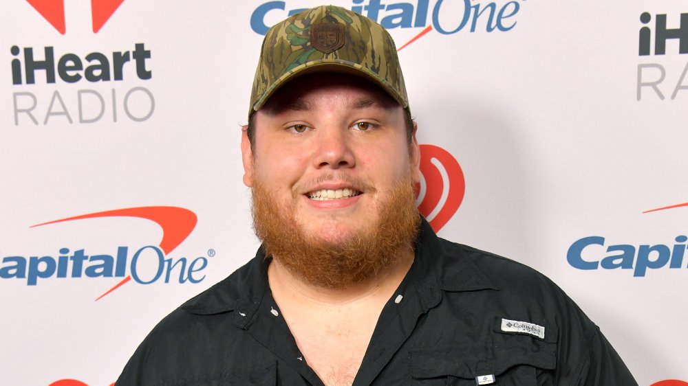 Luke Combs in a black shirt and camo hat, smiling at an iHeartRadio event