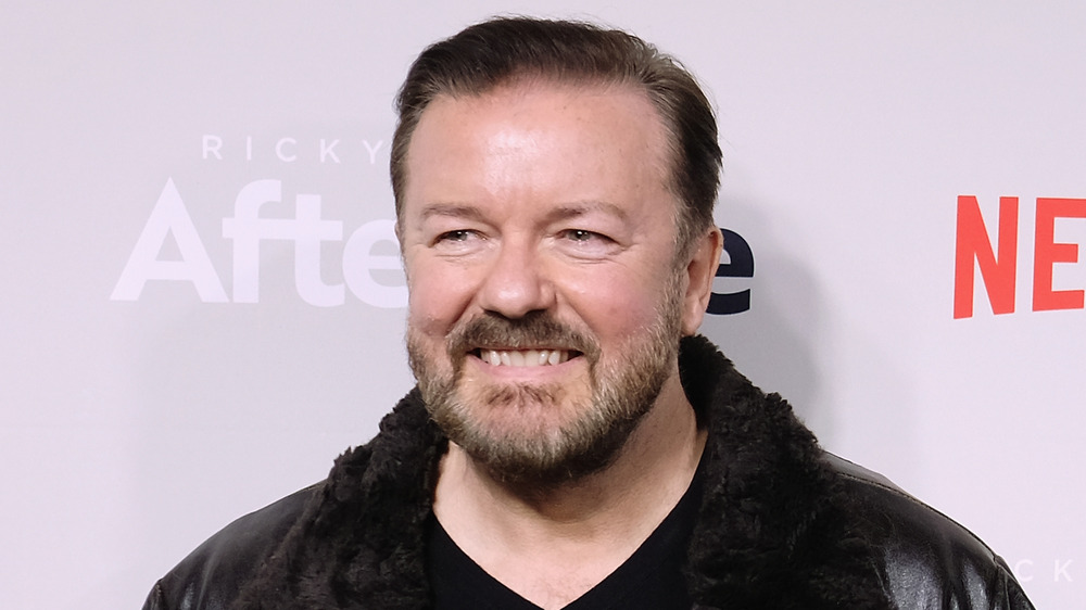 Ricky Gervais smiling in a fur-collared leather jacket