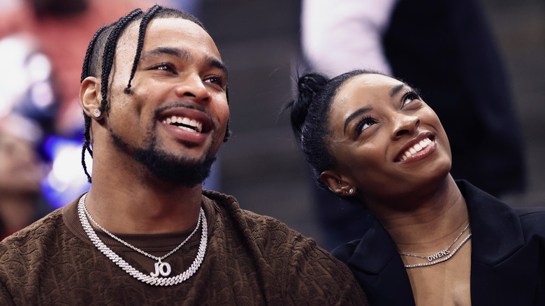 Jonathan Owens wearing a brown shirt and silver chain and Simone Biles wearing a black jacket and silver necklace