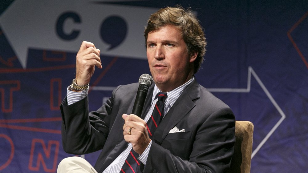 Tucker Carlson in a navy blazer and khaki pants, speaking at an event