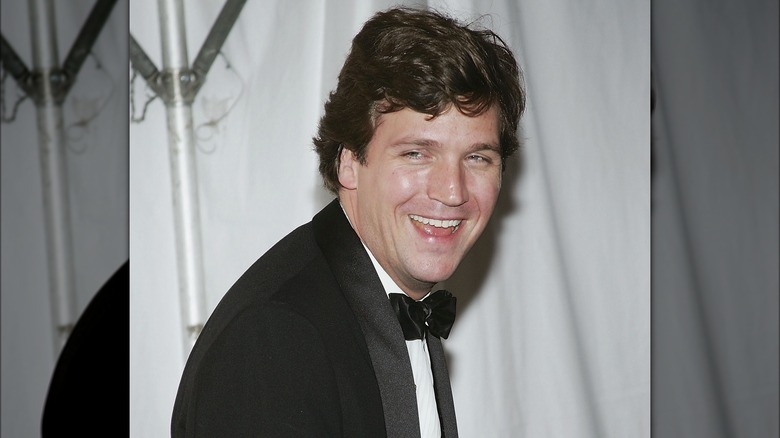 Tucker Carlson in a black suit and bow tie, laughing