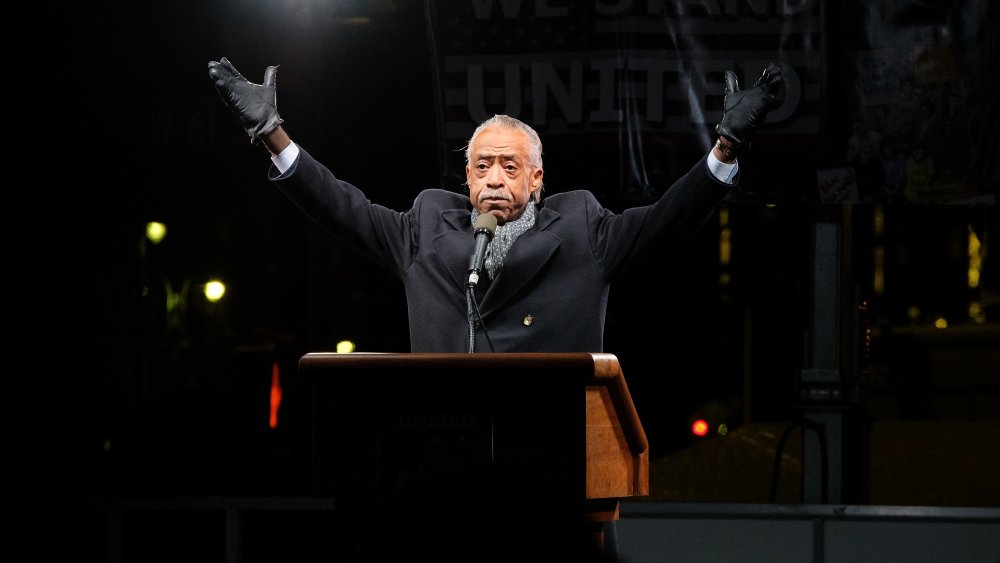 Al Sharpton wearing black gloves with both arms raised 