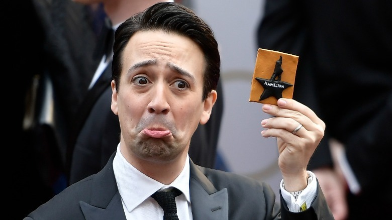 Lin-Manuel Miranda pouts, holding up a Hamilton logo