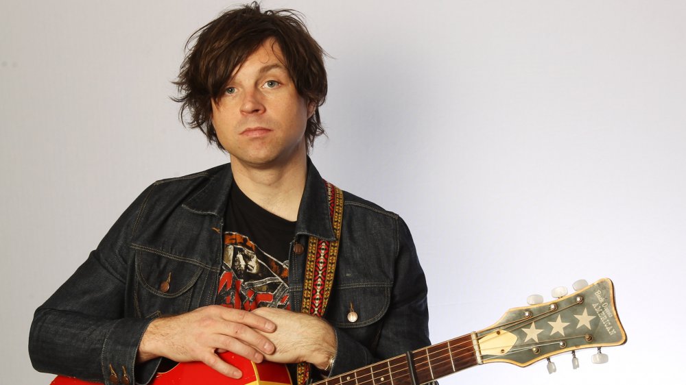 Ryan Adams looking straight ahead with a neutral expression, with his hands folded on top of his guitar