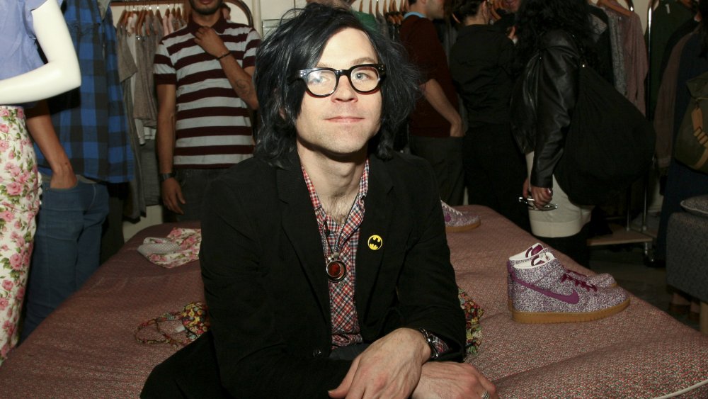 Ryan Adams in a black blazer and red-and-blue plaid shirt, wearing glasses while sitting inside a clothing store