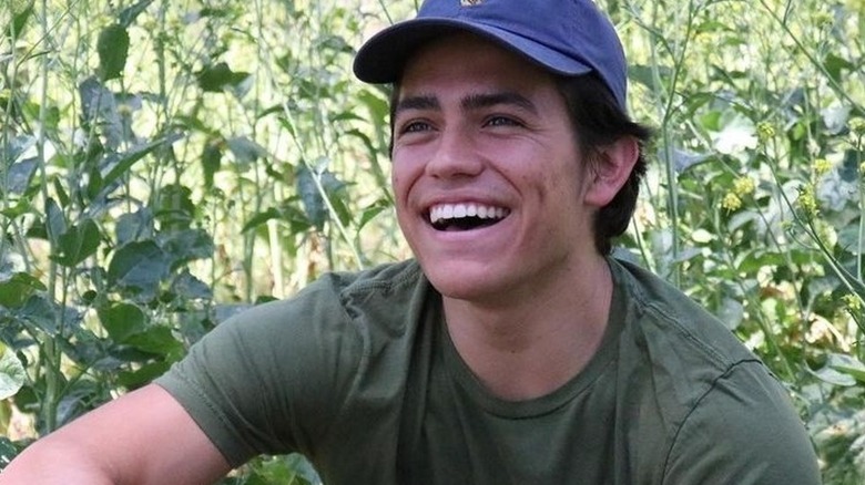 Anthony Barajas smiling while sitting among vegetation with a blue hat on and green shirt