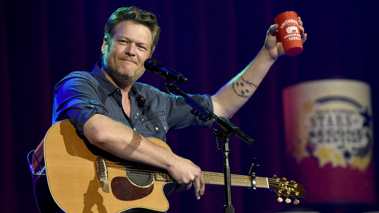 Blake Shelton sits with guitar while extending an arm holding vodka cup.