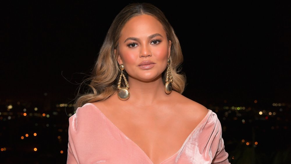 Chrissy Teigen in a pink dress, posing with a neutral expression on a balcony against a city backdrop