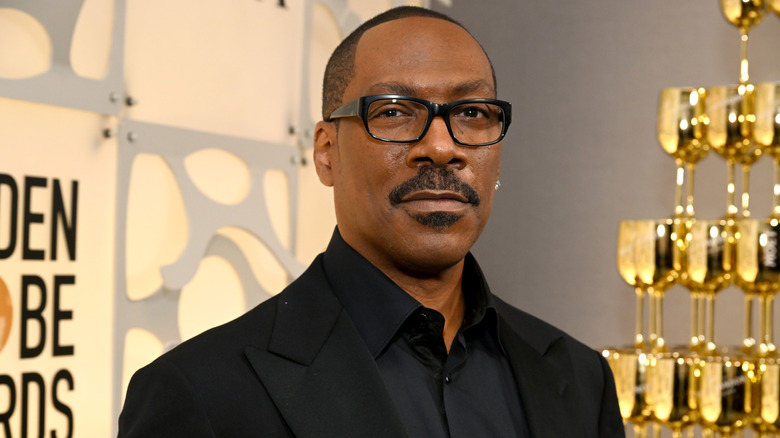 Eddie Murphy wearing glasses, a black button down shirt, and a black formal jacket